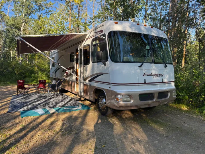 RV rental parked in a treed campsite. Awning extended with camping chairs and outdoor rug on the gravel ground.. Triple E Embassy 2003