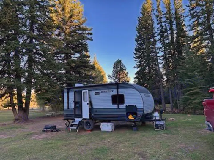 RV rental campsite view: A grey travel trailer sits among tall pine trees under a clear sky, ready for outdoor adventure.. Sunset Park & Rv Inc. 19rb 2025