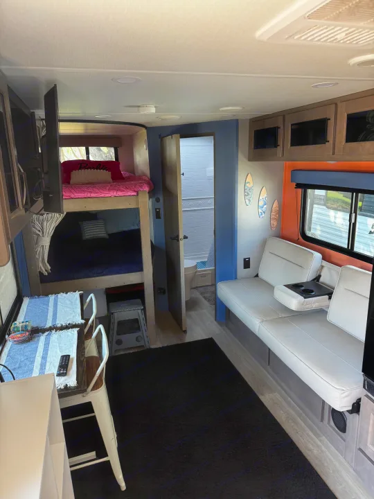 This inviting rv rental interior features bunk beds, a dining area, and a comfortable sofa. A bathroom is visible in the back.. Grand Design Transcend Xplor 2024