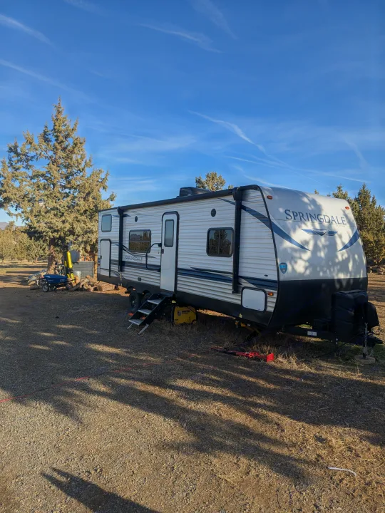 A Springdale RV rental is set up at a scenic campsite with evergreen trees under a bright blue sky, ideal for outdoor travel.. Keystone RV Springdale 2022