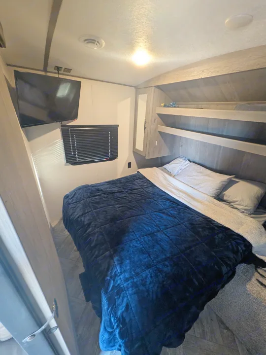 Cozy RV rental bedroom with dark blue bed, mounted TV, window, and storage shelves. Perfect for a comfortable travel stay.. Keystone RV Springdale 2022
