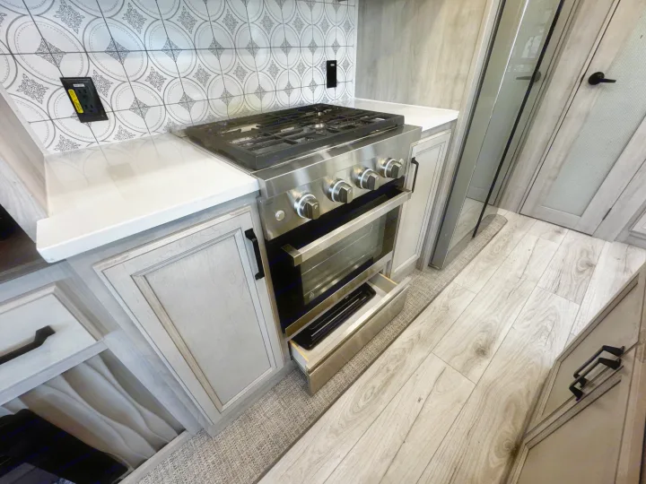 Modern RV rental kitchen with stainless steel stove, patterned backsplash, white countertop, and light wood details.. Keystone RV Montana High Country 2024