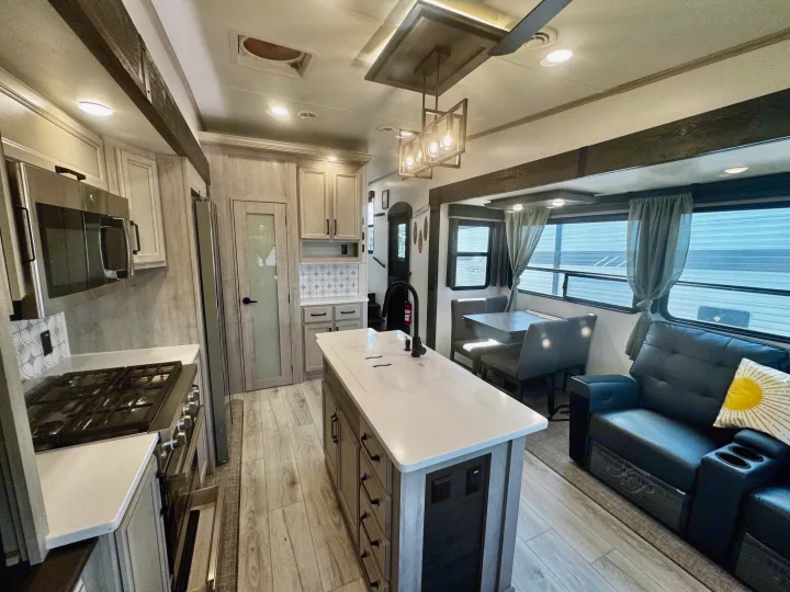 This RV rental offers a bright, modern interior, showing the kitchen island with sink, dining nook, and a black leather sofa.. Keystone RV Montana High Country 2024