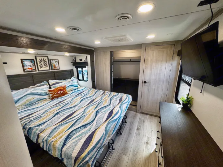 Modern RV rental bedroom: bed with wavy bedding, built-in closet, dresser, and TV. Light wood floors, recessed lighting for comfort.. Keystone RV Montana High Country 2024