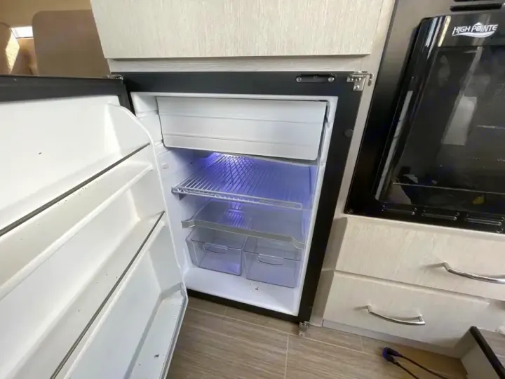 Inside an RV rental, an open compact refrigerator with a small freezer, next to a microwave oven and storage drawers.. Thor Rize 2022