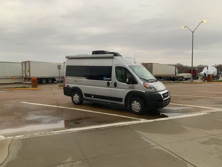 Silver Ram ProMaster camper van, an ideal RV rental, parked at a truck stop under an overcast sky with puddles.. Thor Rize 2022