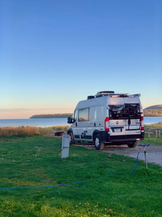 Waterfront RV rental setup: a modern camper van parked at a scenic site with hookups, by a calm lake.. Thor Rize 2022
