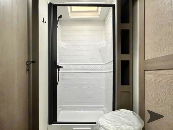 Inside a clean RV rental bathroom: a white shower with black trim, a skylight, and textured walls. Hooks nearby for towels.. Grand Design Transcend 2026
