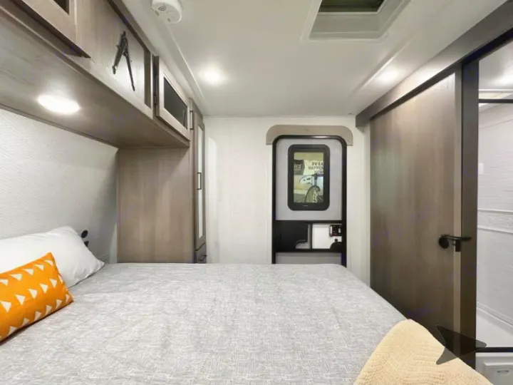 Inside this modern RV rental bedroom: a comfy bed, overhead storage, and entry door, designed for compact comfort.. Grand Design Transcend 2026