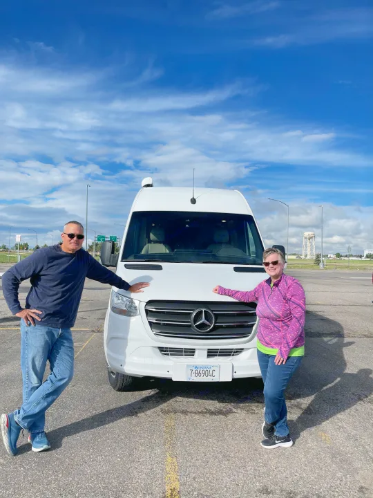 Two people smile proudly with their white Mercedes-Benz Sprinter RV rental, Montana license plate 7-86904C.. Coachmen Galleria 2021