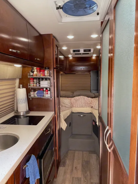 This RV rental interior showcases a compact kitchen with a sink, cooktop, and spice rack, leading to a cozy bedroom area.. Coachmen Galleria 2021