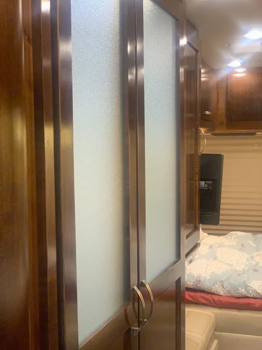 Cozy RV rental interior: Wooden cabinets with frosted glass and curved handles, next to a bed with floral bedding & mounted TV.. Coachmen Galleria 2020