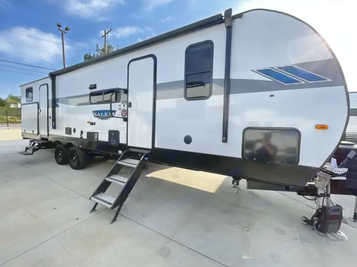 A white travel trailer with black accents & 'Salem' logo, ideal for an RV rental. Steps deployed under a sunny sky.. Forest River Salem Platinum 2024