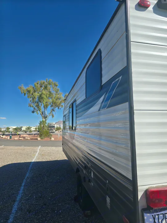 A modern RV rental with striped siding and tinted windows, parked on a gravel lot under a clear blue sky, next to a large tree.. Forest River Salem Cruise Lite 2024