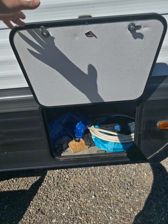 A hand opens an RV rental's exterior storage compartment, revealing essential camping gear like hoses and tarps, with a prominent hand shadow.. Forest River Salem Cruise Lite 2024