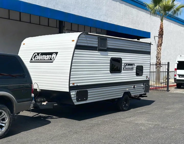 A white Coleman travel trailer, an RV rental, is hitched to a black SUV in a sunny parking lot.. Coleman Lantern 17B 2025