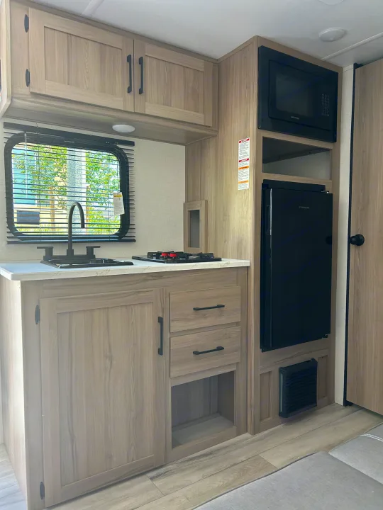 Modern rv rental kitchen featuring light wood cabinets, sink, two-burner stove, microwave, and a black compact fridge.. Coleman Lantern 17B 2025