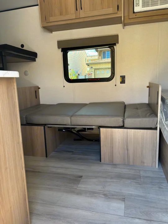 Light RV rental interior features a gray convertible dinette/bed, overhead cabinets, side bunk, window, and wood-look floor.. Coleman Lantern 17B 2025