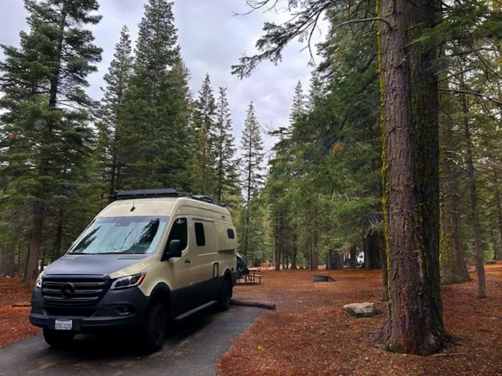 A stylish beige and gray Sprinter RV rental parked in a tranquil forest campground, surrounded by tall pine trees.. Winnebago Revel 2024