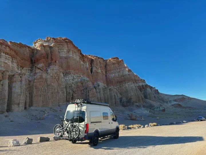 Exploring the desert: An RV rental van with bikes is parked before majestic red rock cliffs under a brilliant blue sky.. Winnebago Revel 2024