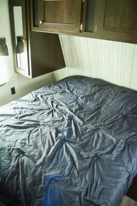 Inside this RV rental's compact bedroom: a rumpled dark gray bed with built-in wood cabinets above for travel accommodation.. Heartland RVs Pioneer 2019