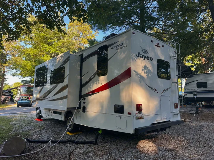 A white Jayco Redhawk RV rental at a gravel campsite. Slide-outs are extended, utilities connected, and trees shade the scene.. Jayco Redhawk 2018