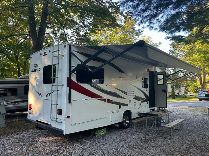 Relaxing RV rental on a gravel campsite under lush trees. The Jayco motorhome has its awning extended and a picnic table nearby.. Jayco Redhawk 2018