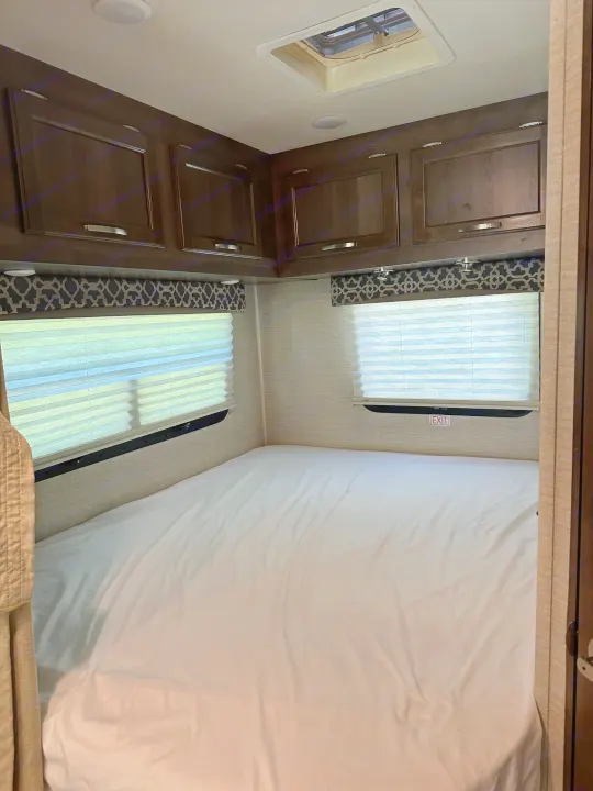 This RV rental's cozy bedroom features a white bed, dark overhead cabinets, and windows with blinds for comfort.. Jayco Redhawk 2018