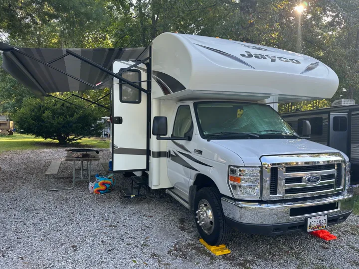 White Jayco rv rental with an extended awning at a gravel campsite, a picnic table nearby under sun-dappled trees.. Jayco Redhawk 2018
