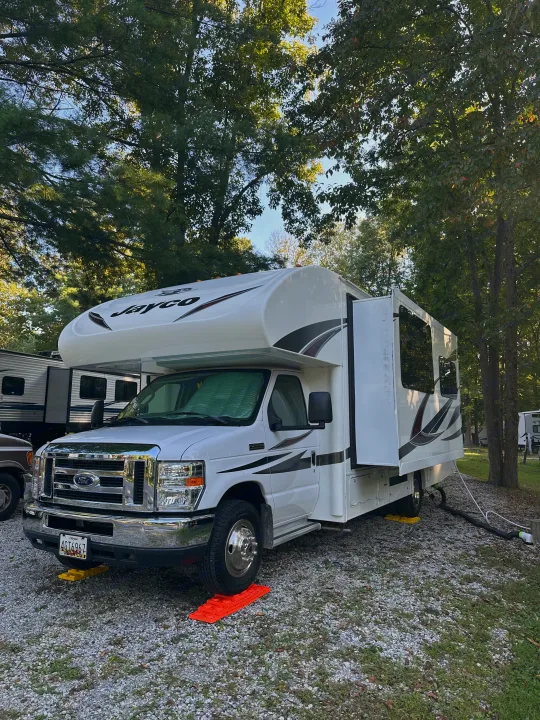 A white Jayco RV rental parked at a tree-filled campsite, showing leveling blocks and utility hookups for camping.. Jayco Redhawk 2018