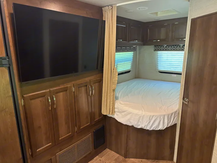 RV rental bedroom interior: large TV, bed with white sheets, overhead cabinets, and windows with blinds.. Jayco Redhawk 2018