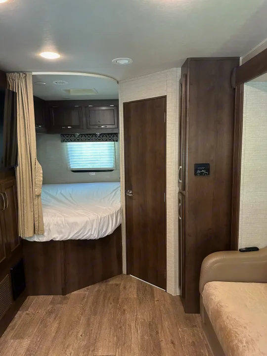 Inside an RV rental: a compact bedroom with a window, dark wood cabinets, and a comfy sofa in the main living area.. Jayco Redhawk 2018