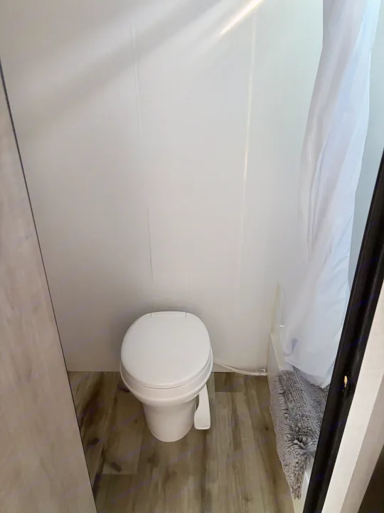 An RV rental's compact bathroom features a white toilet, shower curtain, wood-grain floor, and sunlight on white walls.. Dutchmen Kodiak Cub 2024