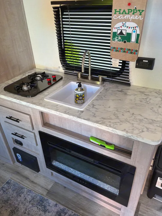 This RV rental kitchen features a two-burner stove, sink, and electric fireplace. A 'Happy Camper' towel hangs nearby.. Dutchmen Kodiak Cub 2024