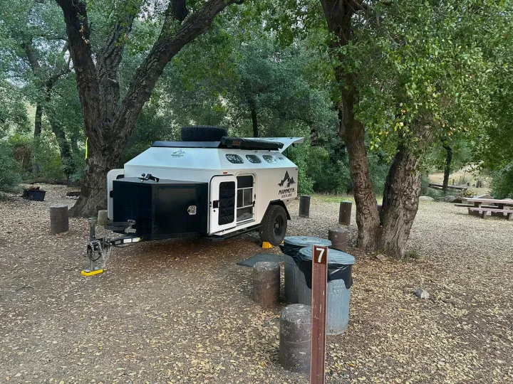 A white overlanding RV rental trailer is parked in a shaded campsite, surrounded by green trees, with trash cans and a picnic table.. Mammoth Overland HV 2022