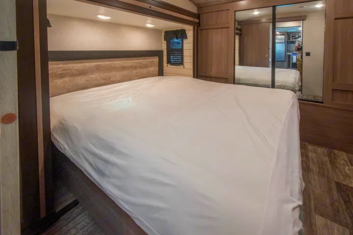Spacious RV rental bedroom featuring a white bed, wood-textured headboard, window, and mirrored closet reflecting interior space.. Crossroads RV Sunset Trail 2022