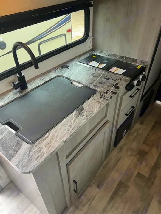 Modern RV rental kitchen with marble-look counter, black faucet, sink, covered stovetop, and light wood grain cabinets for a stylish trip.. Dutchmen Kodiak Cub 2022