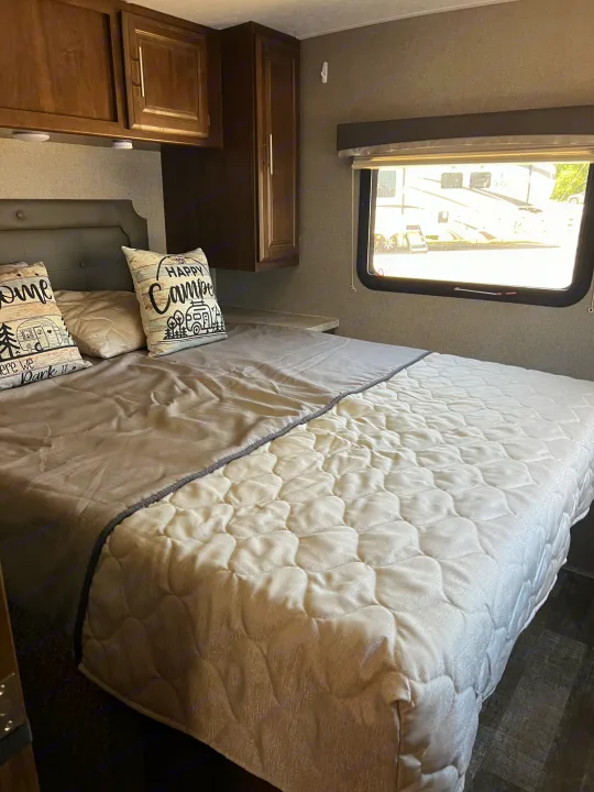 Cozy RV rental bedroom with a bed, "Happy Camper" pillows, wooden cabinets, and a bright window view.. Forest River Rockwood Mini Lite 2020