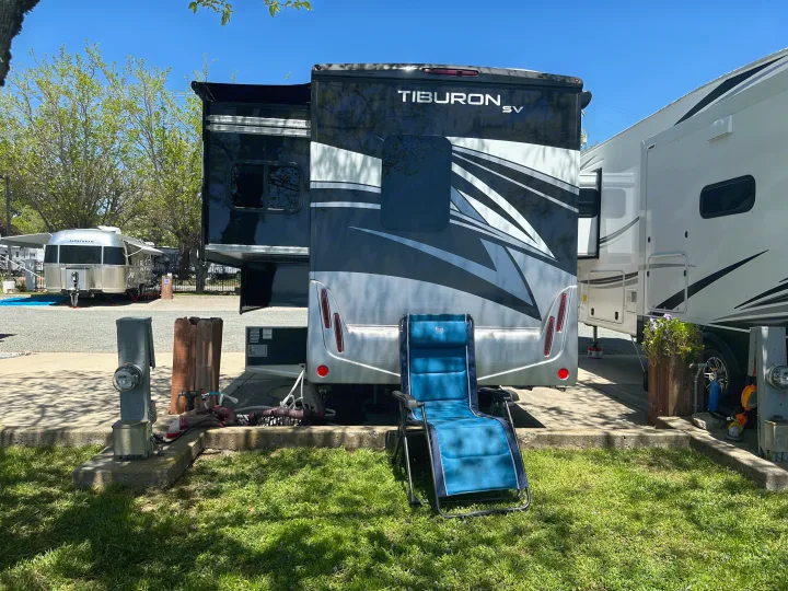 Black & silver Tiburon SV RV rental in a sunny campground, with a blue reclining chair, utility hookups, and an Airstream nearby.. Thor Tiburon SV 2023