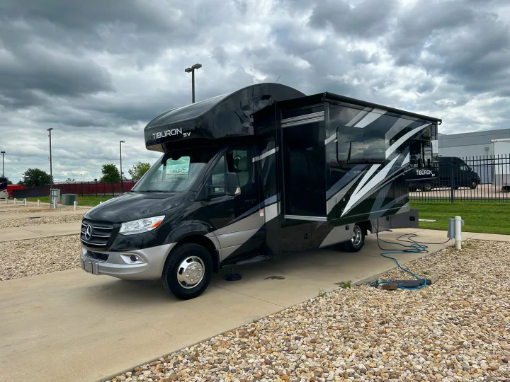 This black and silver Tiburon SV RV rental is parked at a campsite on a cloudy day, with its slide-out extended and utilities connected.. Thor Tiburon SV 2023