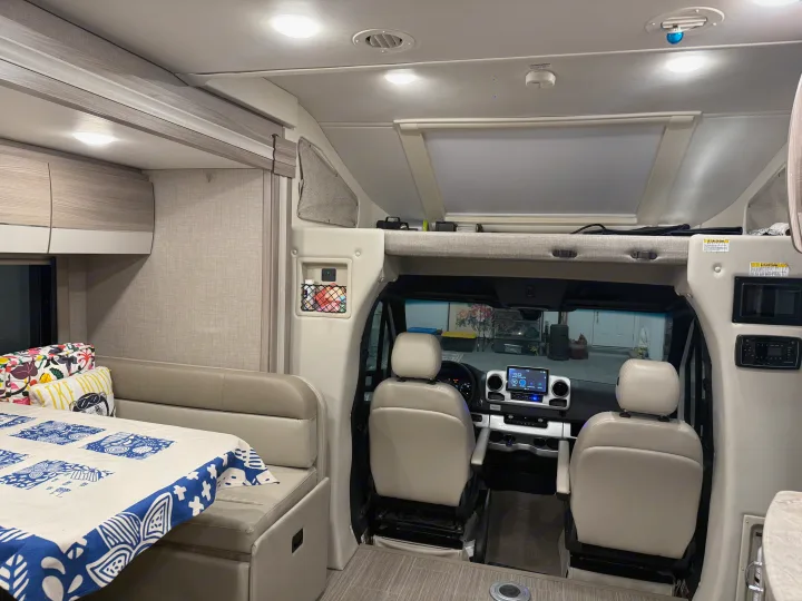 View of a modern RV rental interior showing a dinette with patterned tablecloth, cockpit, and overhead bunk.. Thor Tiburon SV 2023