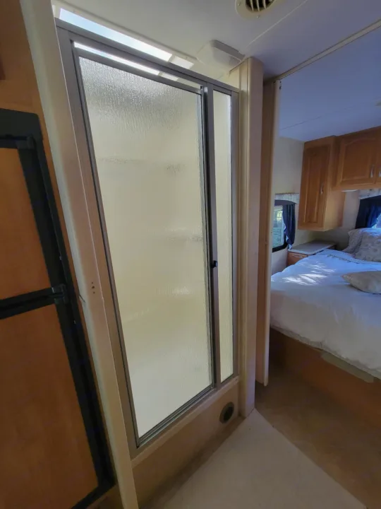 Compact RV rental interior: A frosted shower stall on left, a cozy bedroom with a white bed visible on the right.. Forest River Sunseeker LE 2006