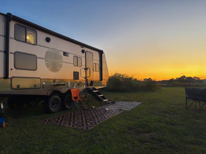 RV rental journey: A 'no-bo' travel trailer parked in a wide grassy field, bathed in warm sunset hues. An outdoor mat and chair await.. Forest River 5NHTRDV27TD211453 2026