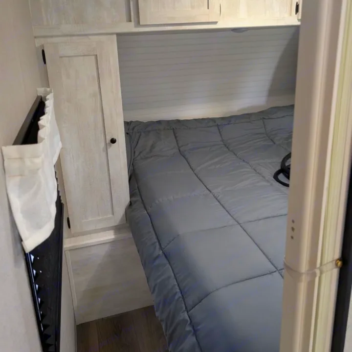 Cozy RV rental bedroom featuring a grey comforter, light wood cabinetry, and a small curtain.. Coachmen I-5 Edition 530QB 2024