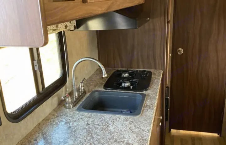 This rv rental kitchen features a compact sink, two-burner stove, range hood, and wooden cabinets, ideal for travel.. Coachmen Clipper 2018