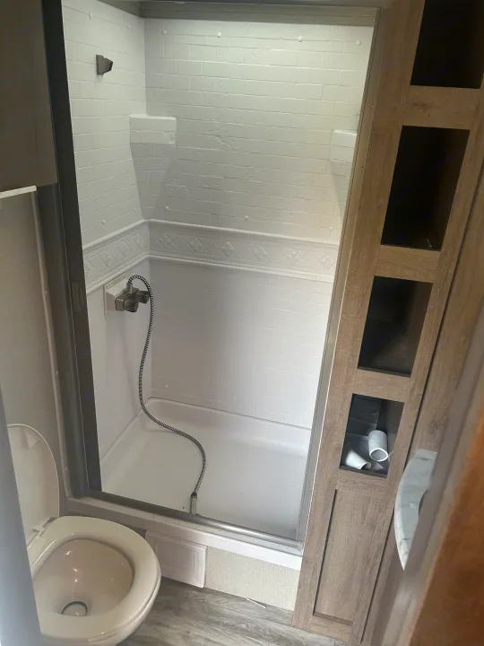 This RV rental bathroom features a compact white shower stall with a handheld showerhead, a toilet, and integrated storage.. Grand Design Imagine 2021