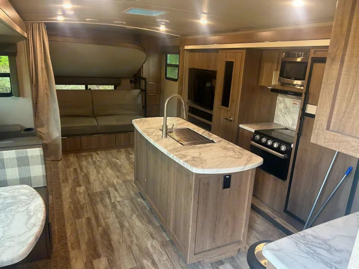View of an RV rental interior with a central kitchen island, oven, microwave, and living area with a sofa and bunk.. Grand Design Imagine 2021