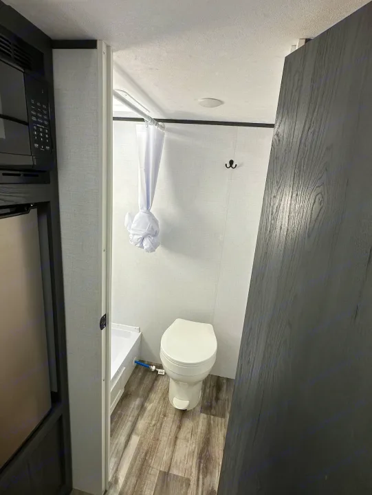 RV rental bathroom features a toilet, shower with curtain, and adjacent microwave/fridge unit in a compact layout.. Dutchmen Other 2024