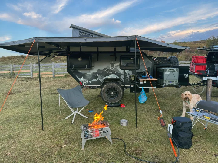 Scenic RV rental setup: overlanding trailer, cozy campfire, happy dog, and outdoor cooking station under an awning.. Mission Overland Summit 2024