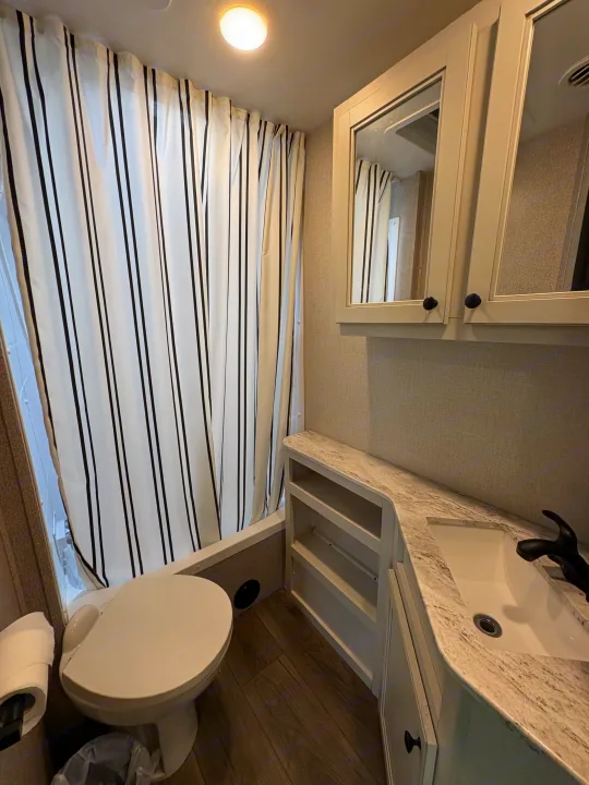 RV rental bathroom: striped shower curtain, mirrored cabinet, vanity with sink & shelving, and toilet.. Highland Ridge RV Range Lite 2021